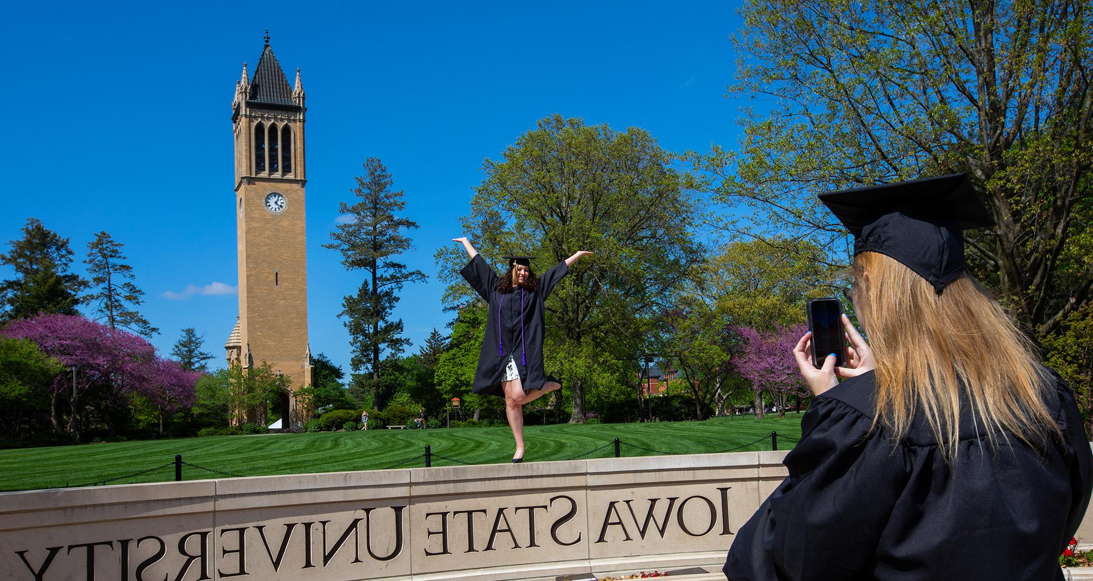 A graduate takes a picture of another graduate on the Iowa State University wall in front of the campanile