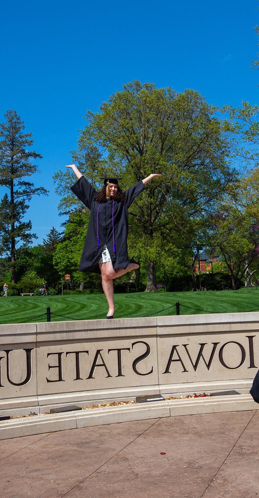 A graduate poses on the Iowa State University wall in front of the campanile